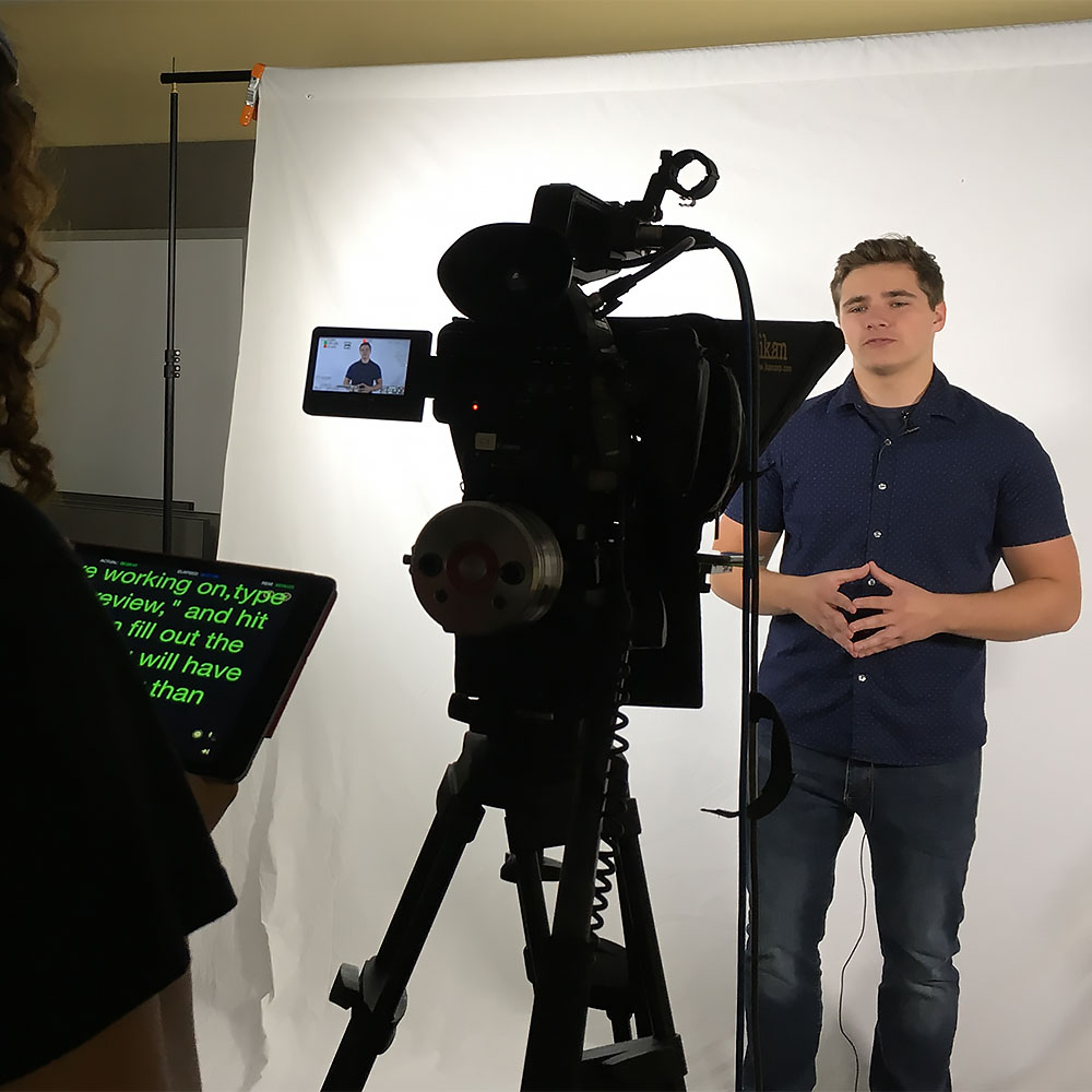 A student stands in front of a video camera while reading a script from a teleprompter.