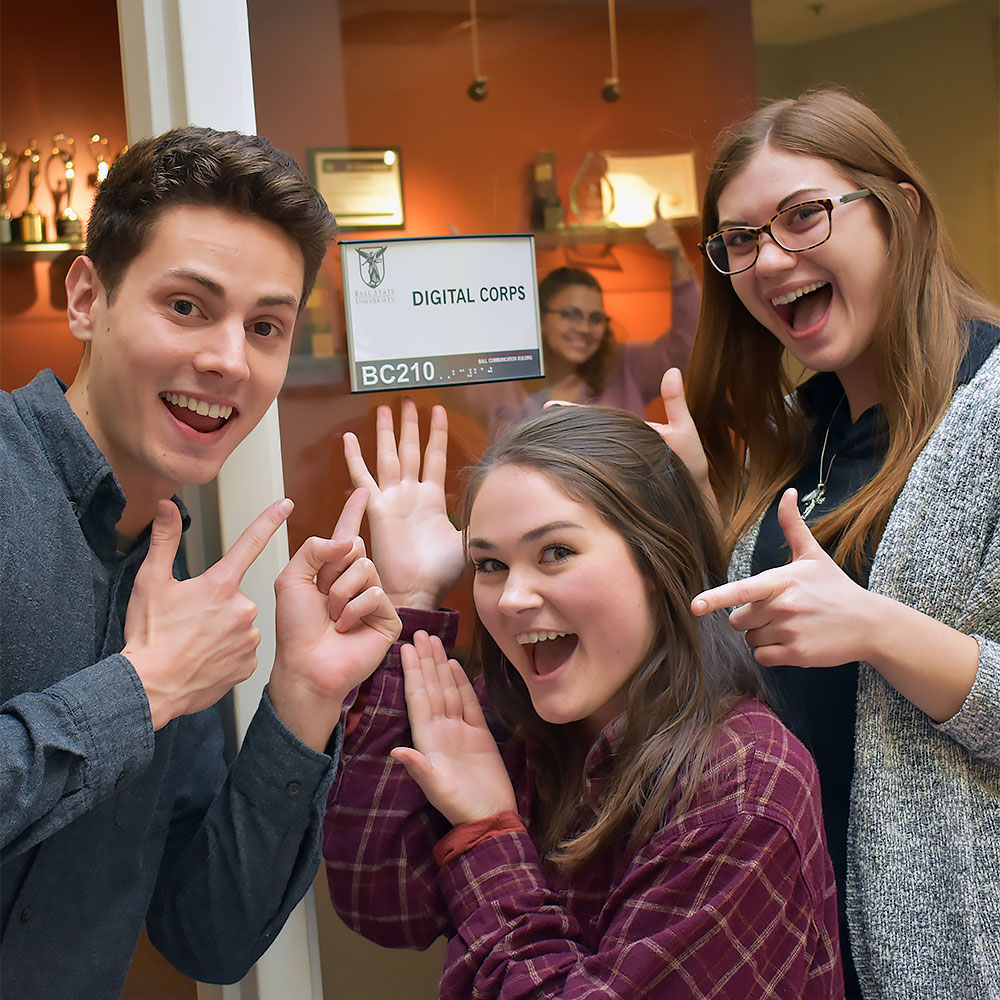 A group of excited college students stand outside an office with a sign that reads “Digital Corps”