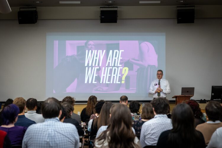Students gathered in a lecture hall being given a presentation. The screen displays the words "Why are we here?"
